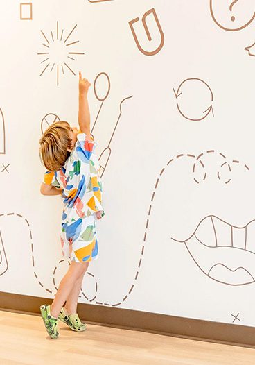 Child pointing at dental-themed wall illustration in pediatric dental office